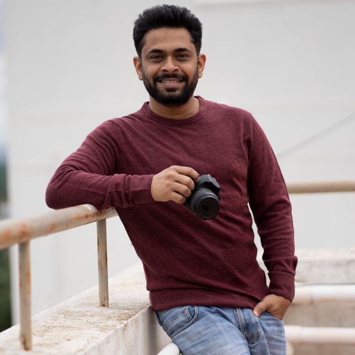 Indian man in casual attire holding a camera, smiling outdoors.